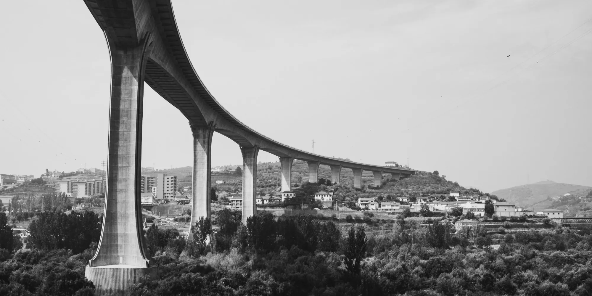 Black-and-white photo of a tall curving concrete highway viaduct on slender piers above a hillside town and terraced slopes.