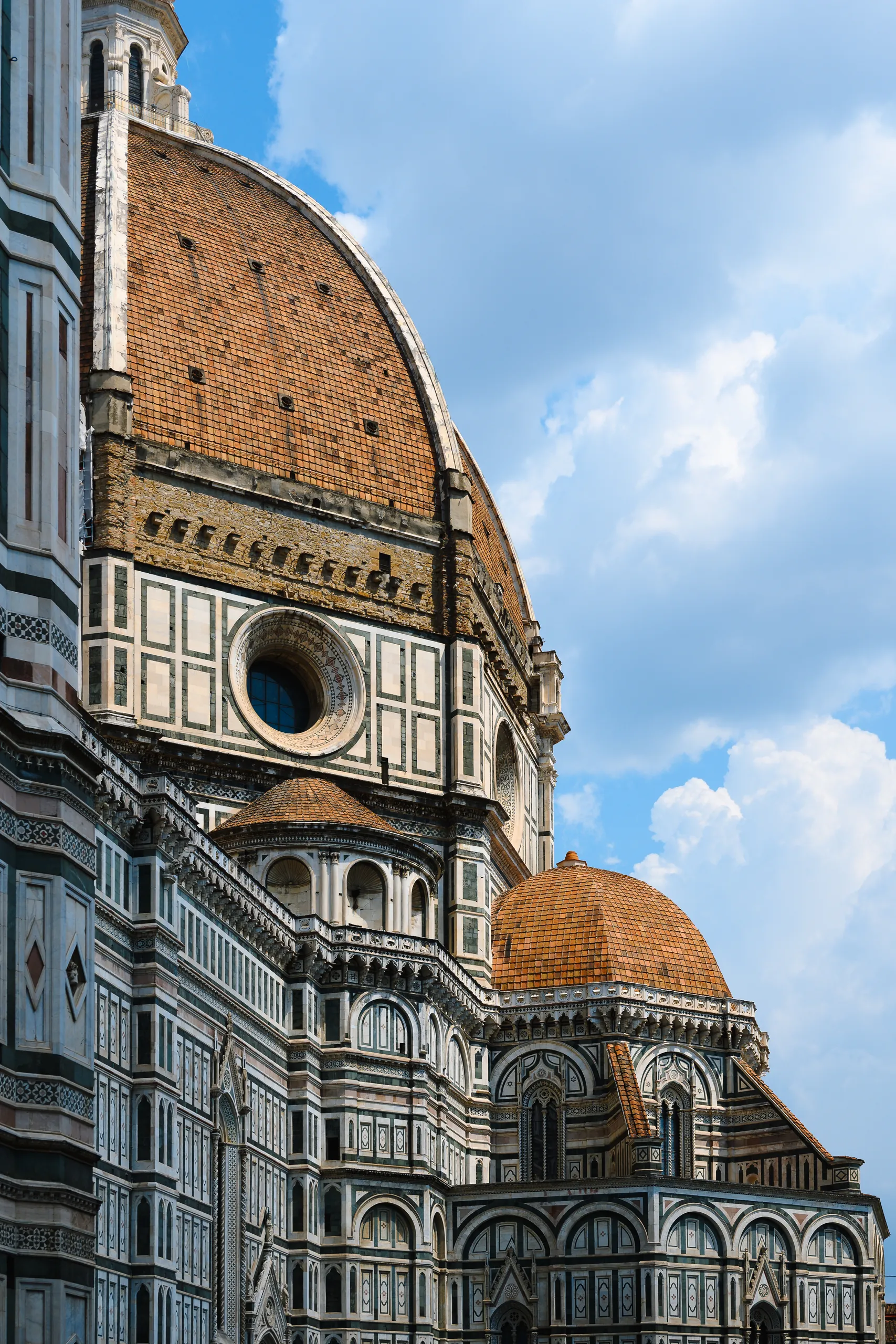 Looking up at the red-tiled dome of Florence Cathedral and its marble facade in white, green and pink, against a blue sky with clouds.