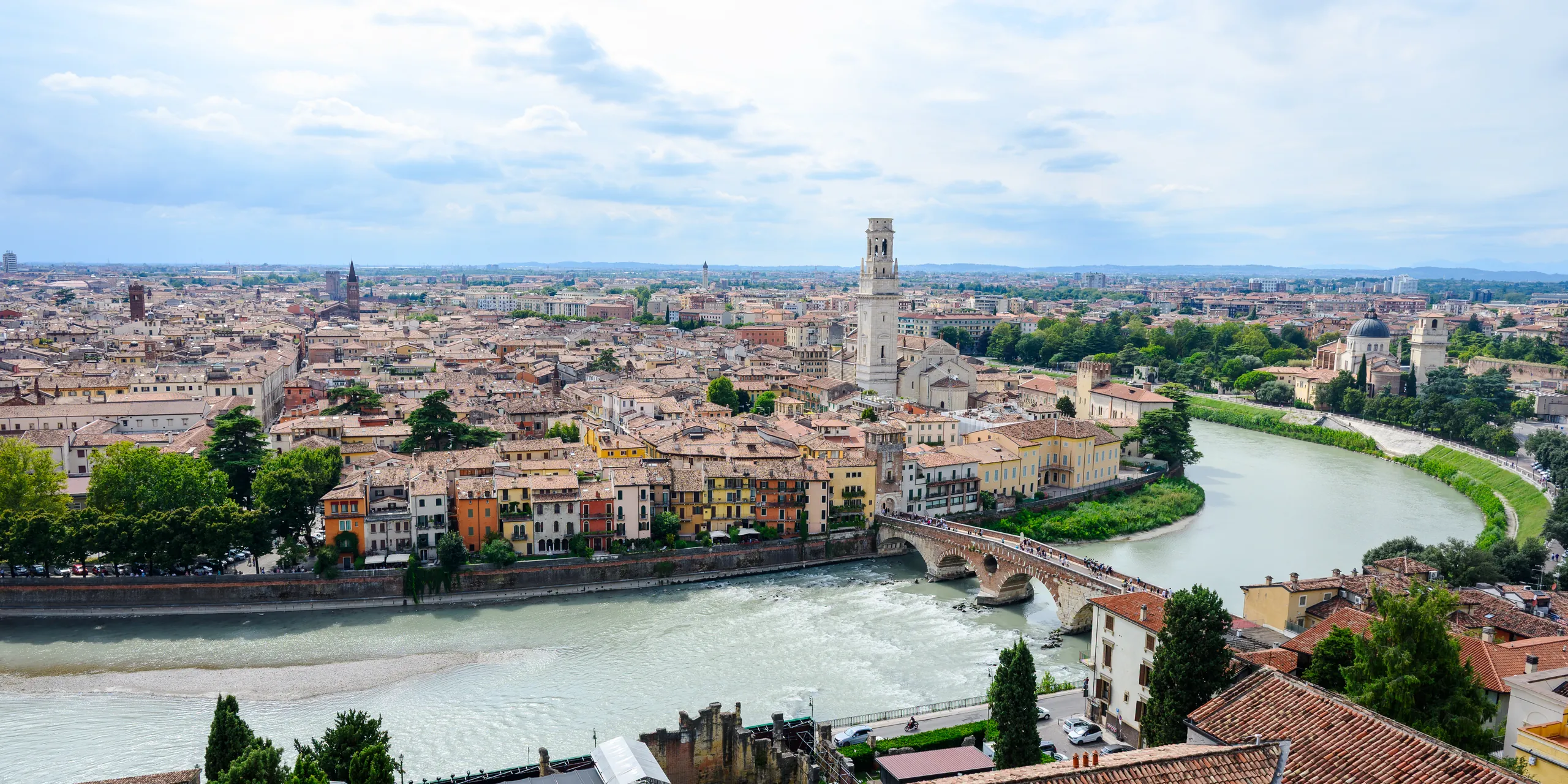 Panorama of Verona's terracotta rooftops and bell towers along a bend of the Adige River, seen from a hilltop.