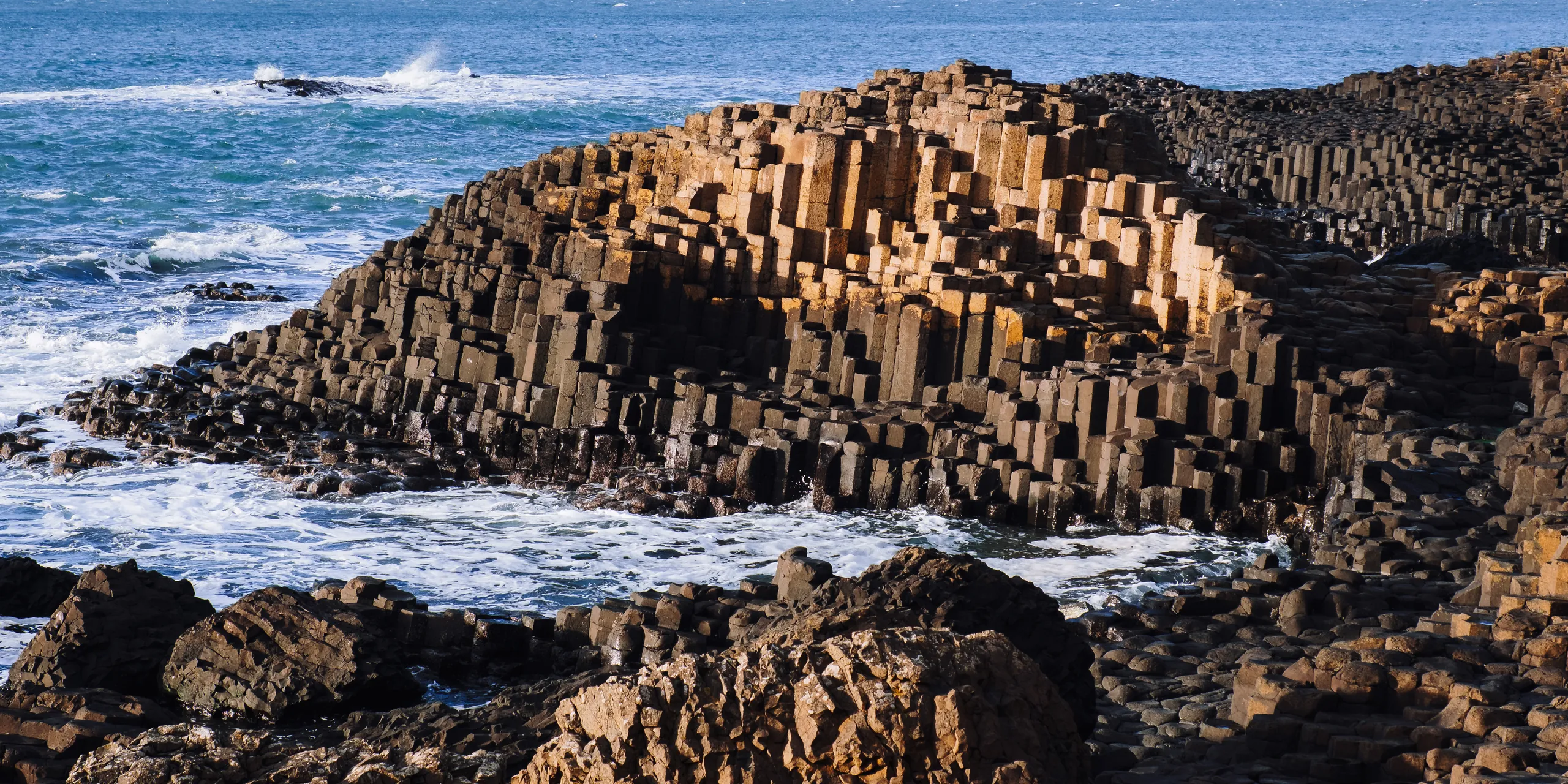 Hexagonal basalt columns stepping down into the sea at the Giant's Causeway, with waves breaking white around the rocks.