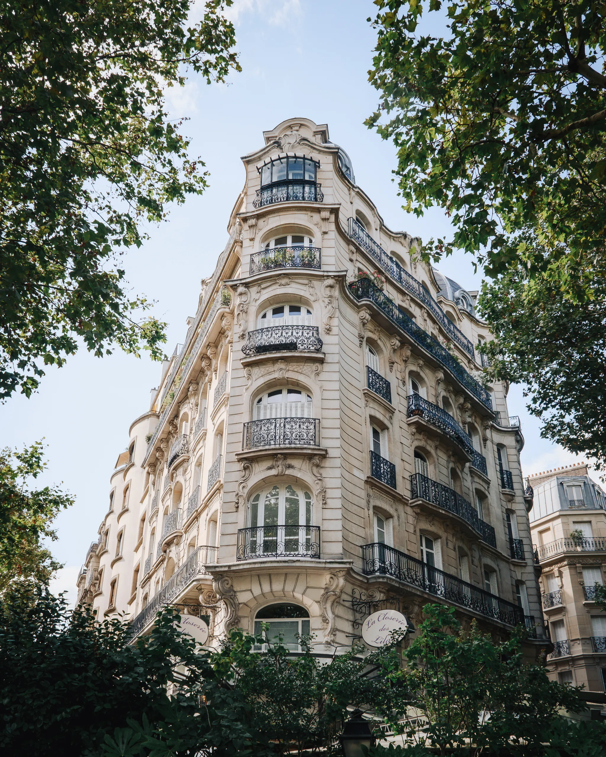 Looking up at an ornate cream stone Parisian apartment building with wrought-iron balconies, framed by green trees and blue sky.