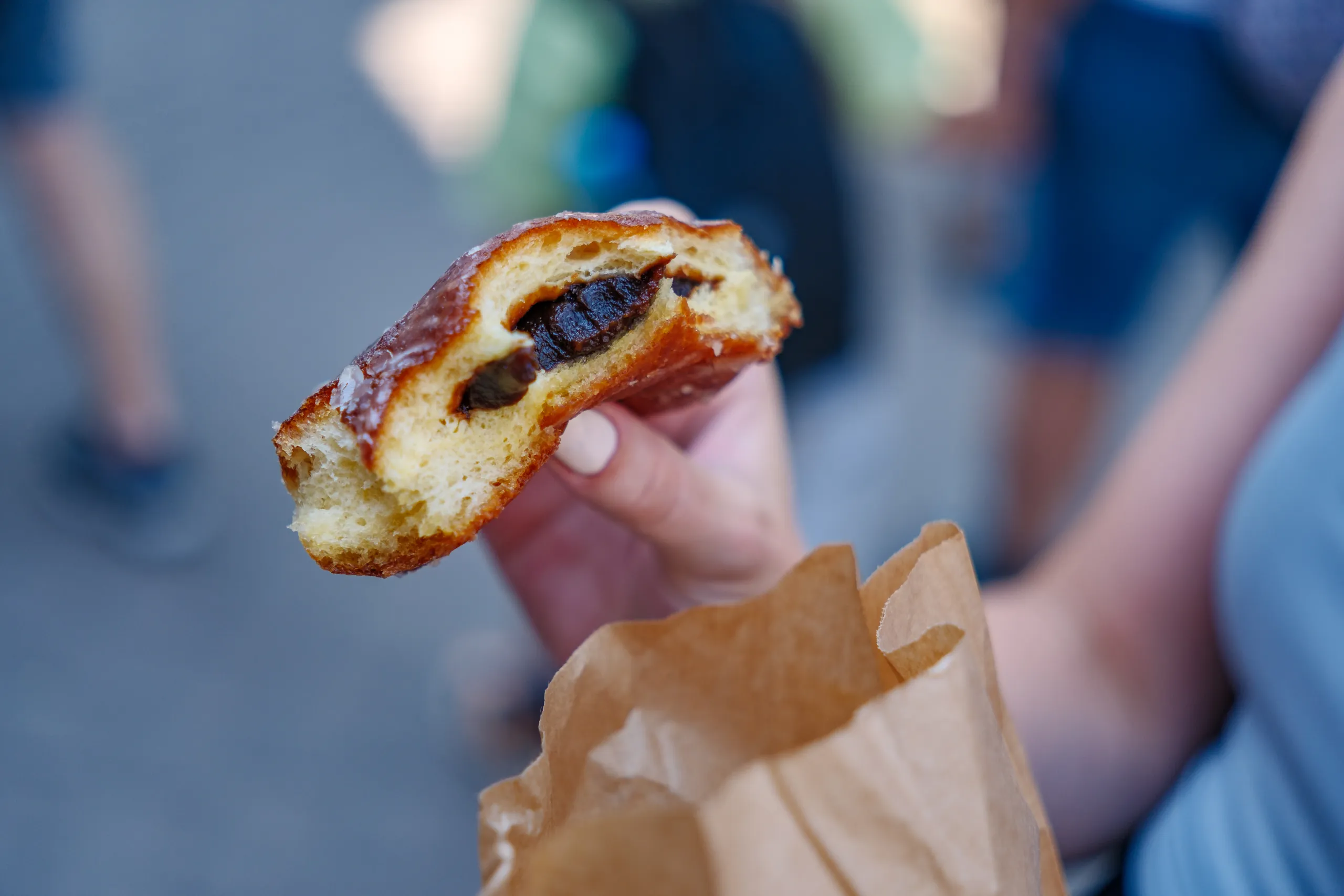 A hand holding a half-eaten pączek doughnut with dark plum filling above a paper bag, on a blurred street.