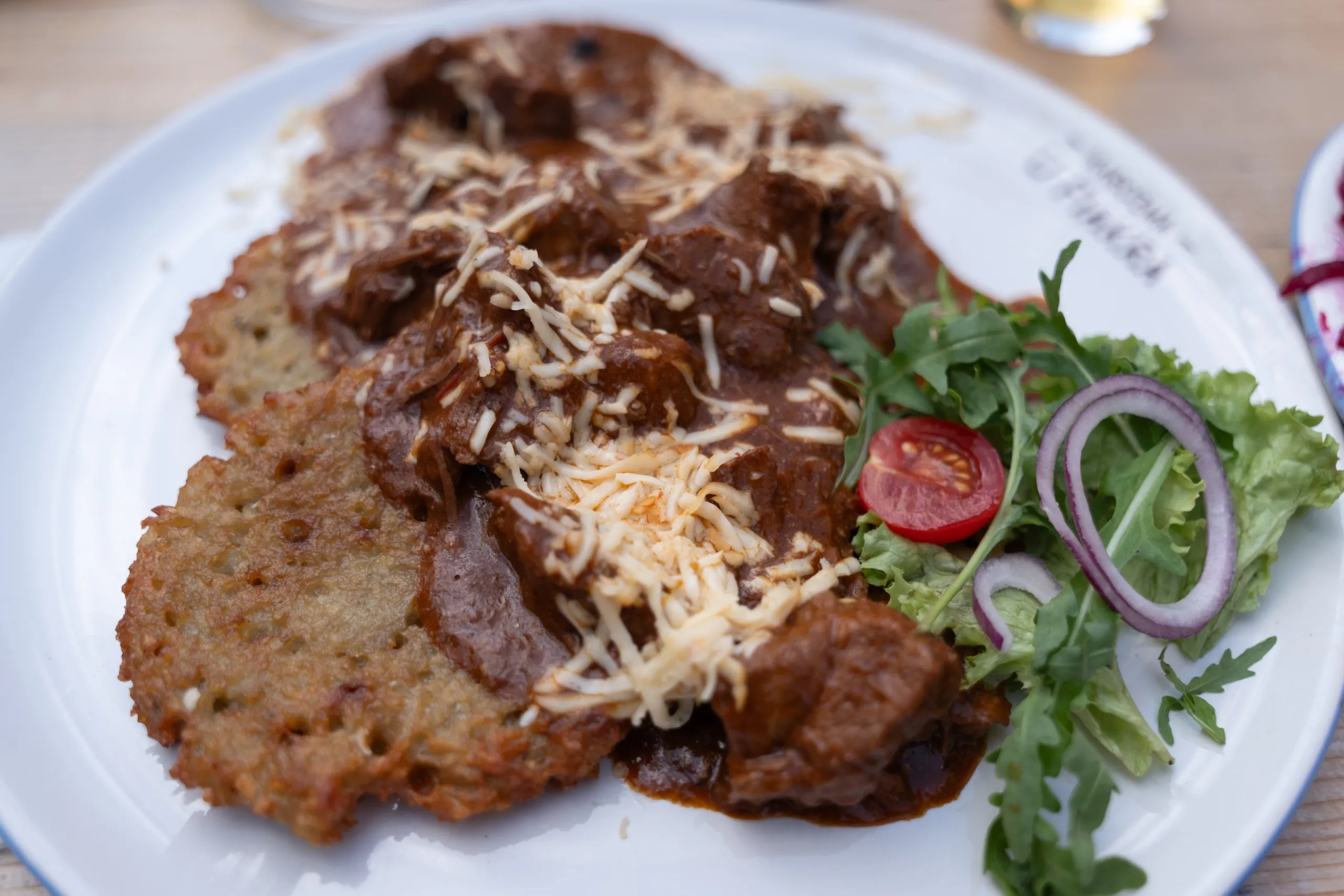 Potato pancakes covered in beef goulash and grated cheese on a white plate, with a side salad of greens, red onion and cherry tomato.