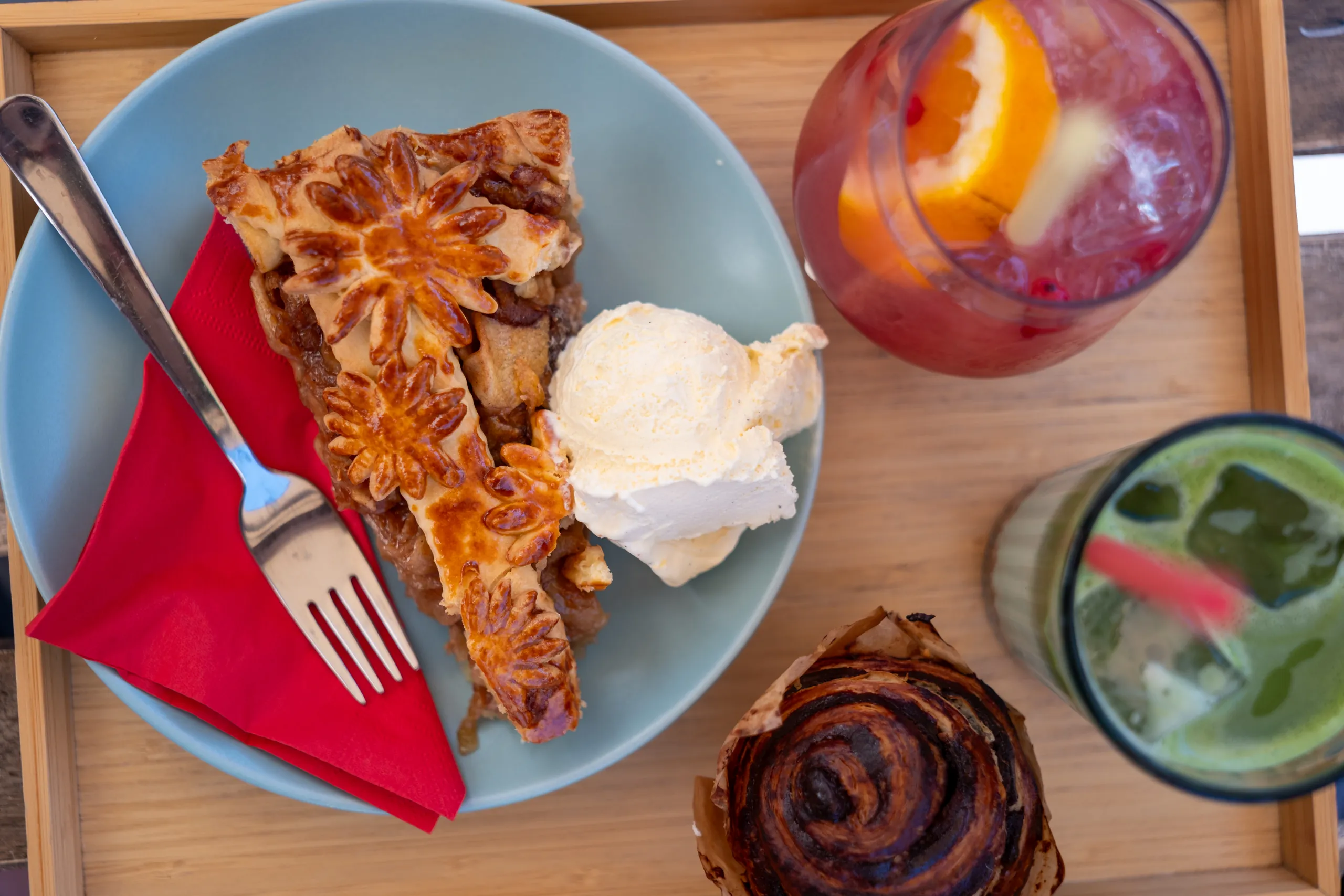 Overhead view of a wooden tray with a slice of lattice apple pie and ice cream, a cinnamon roll, and two iced drinks with orange and mint.