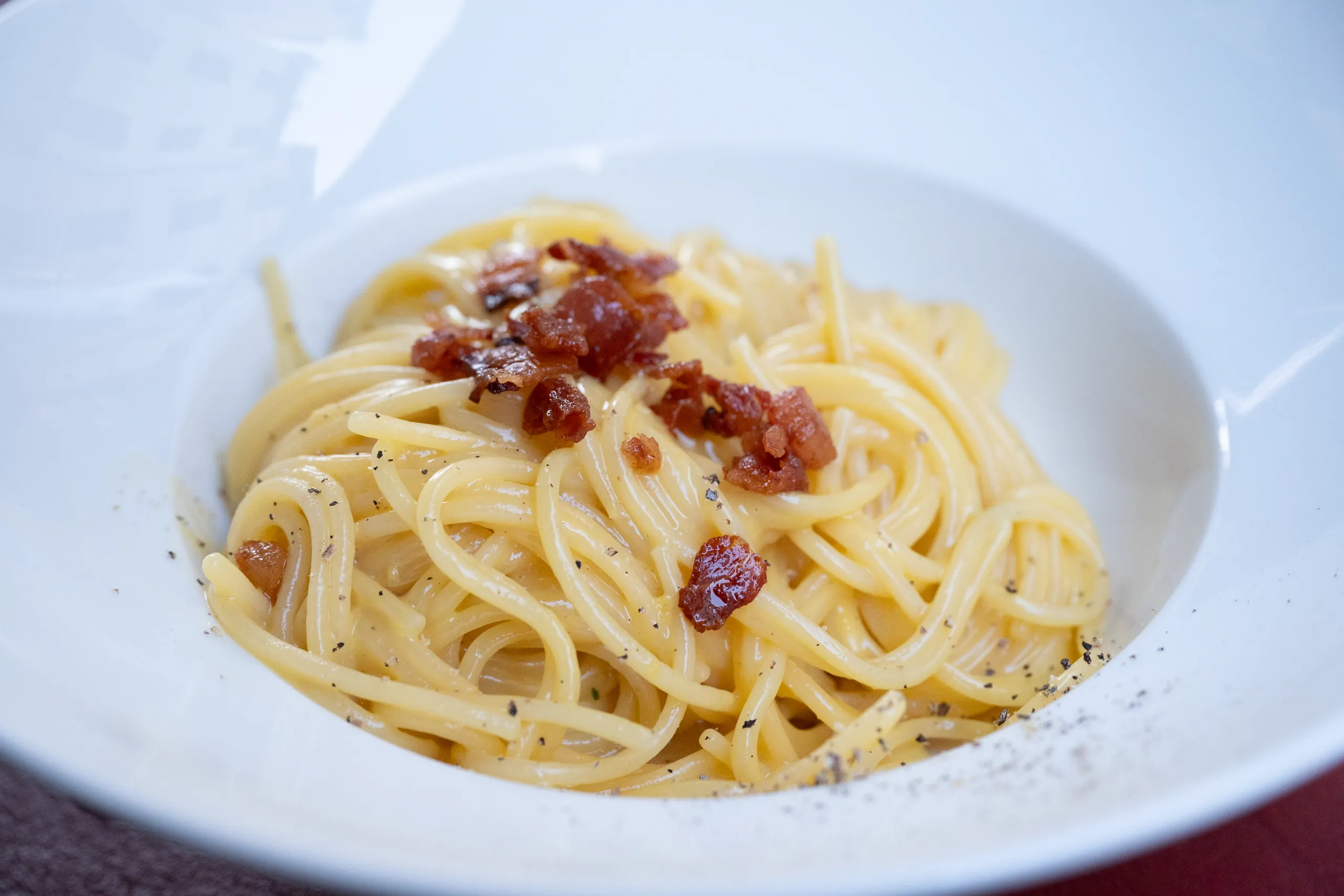 Close-up of spaghetti carbonara topped with crisp pieces of guanciale and black pepper in a white bowl.
