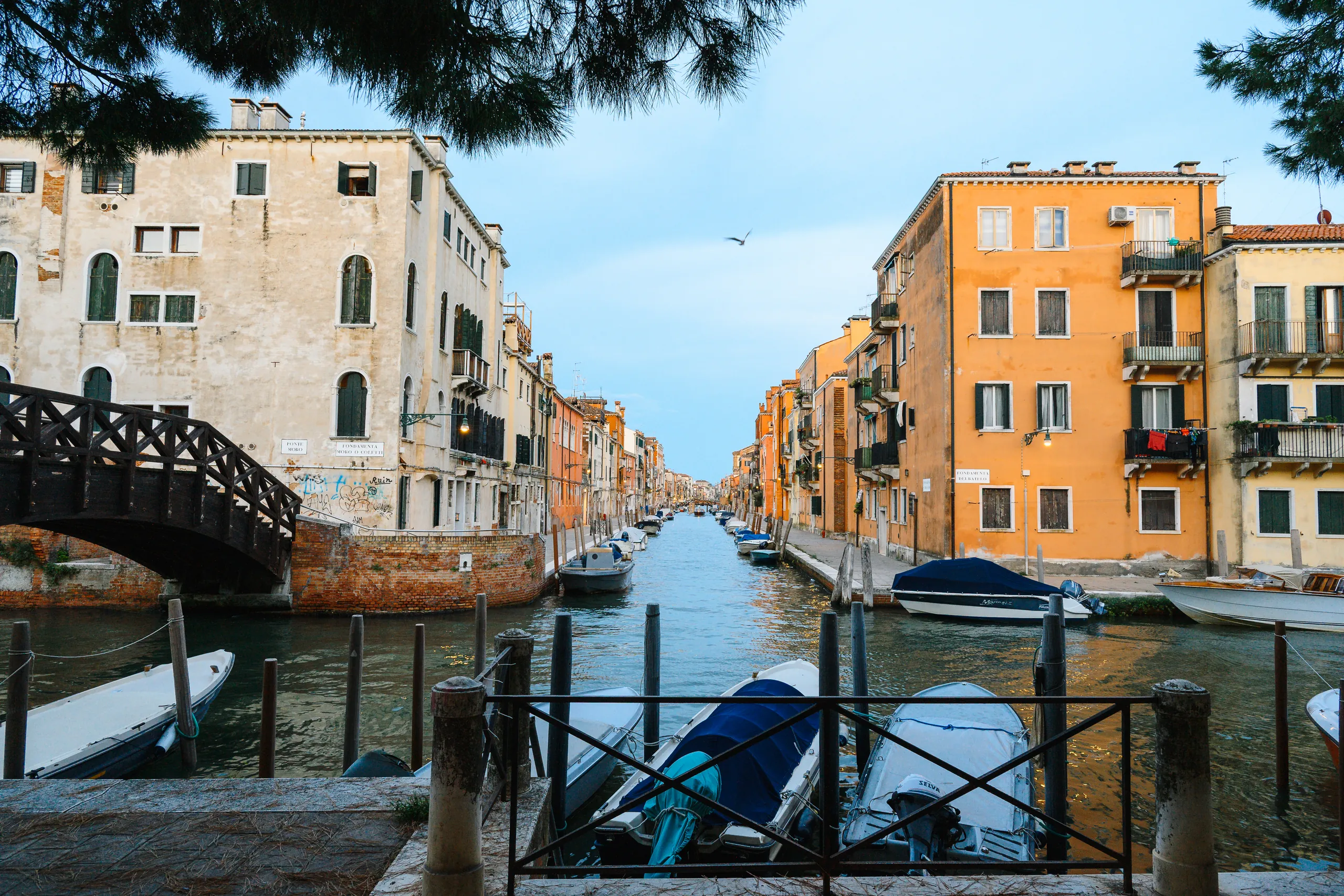 A quiet Venetian canal lined with pastel and orange buildings, a wooden footbridge on the left and small boats moored along the water.