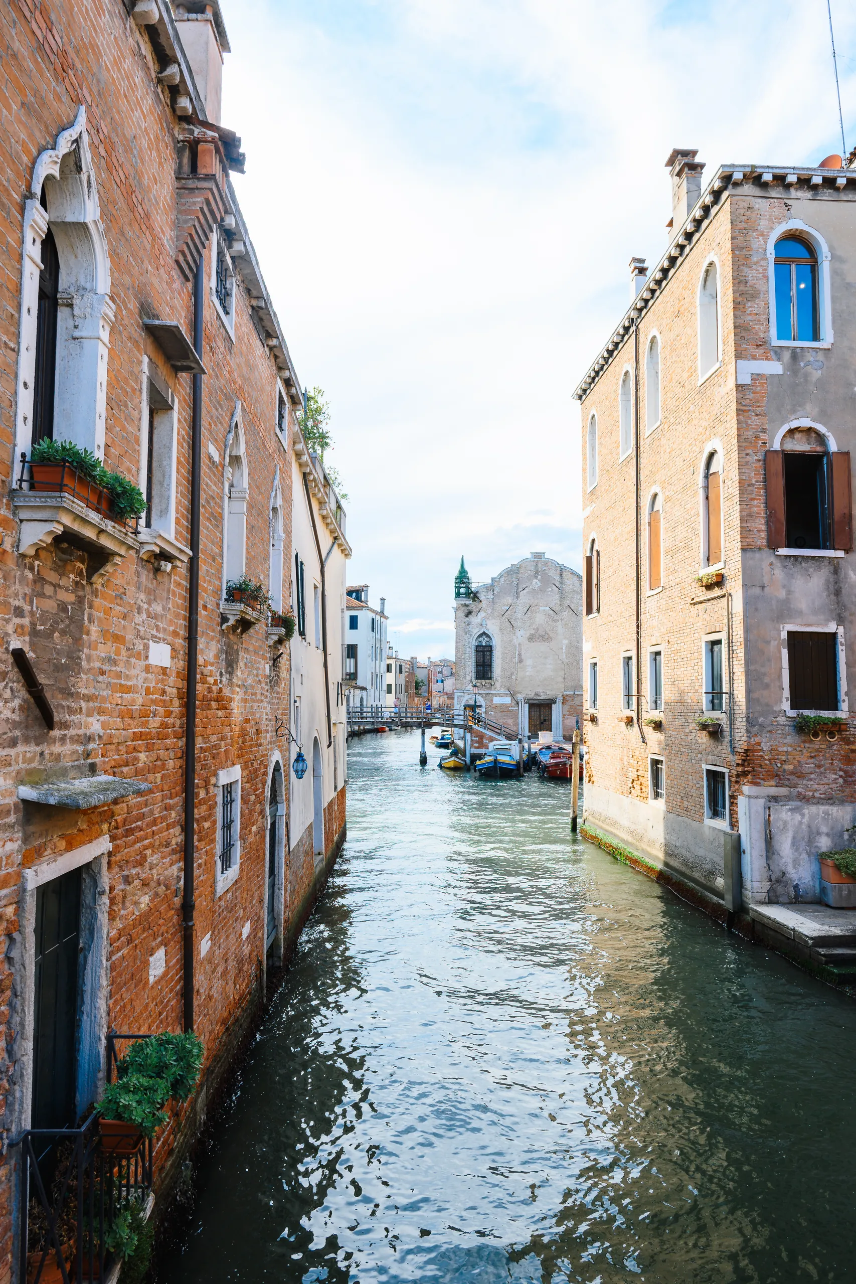 Vertical view down a narrow canal between brick buildings with flower boxes, leading to a church facade at the far end.