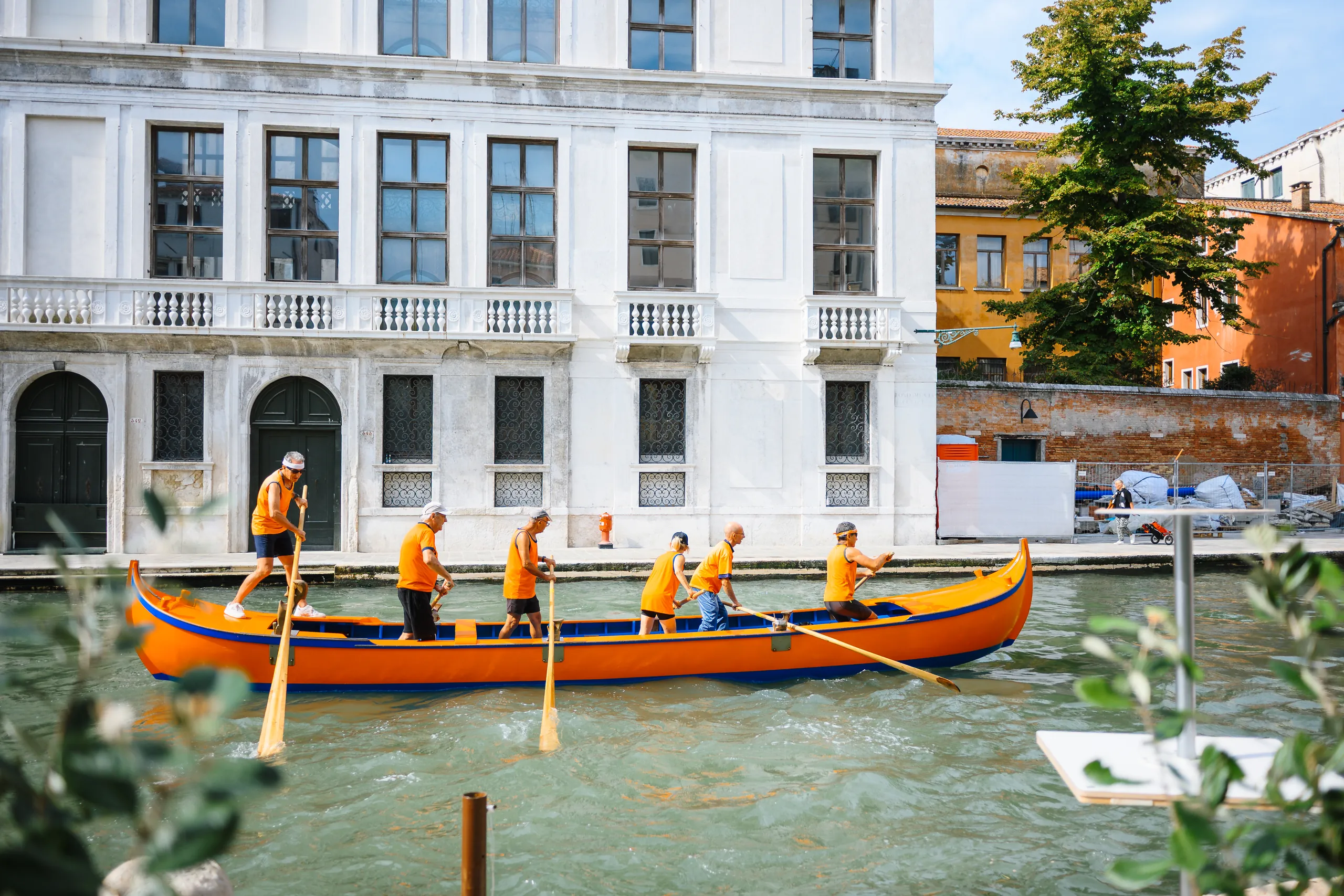 Six rowers in orange shirts standing and rowing a long orange boat along a Venetian canal past a white palazzo.
