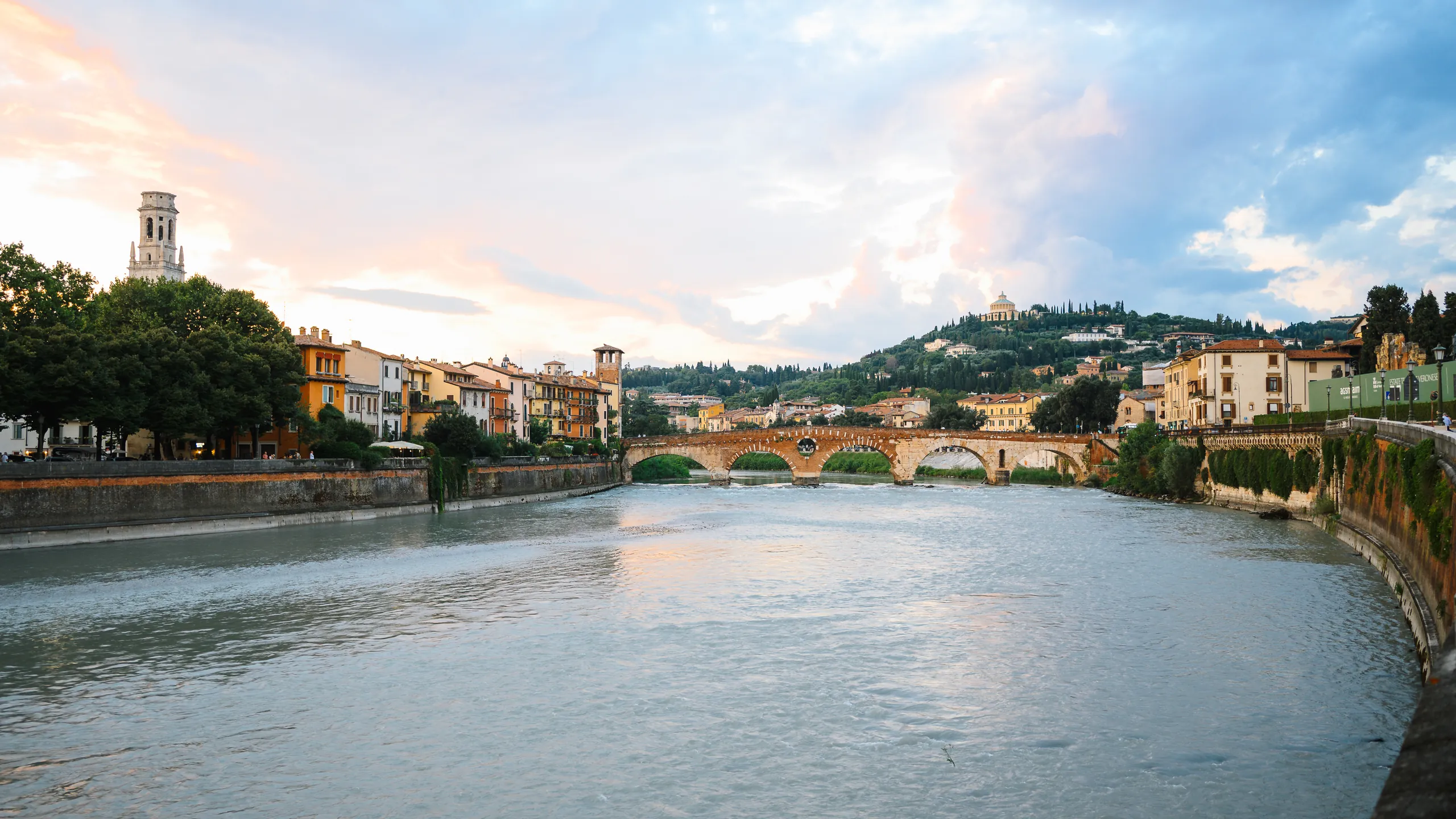 The Adige River curving through Verona at sunset, with a stone bridge, riverside buildings and a hill under pink and blue clouds.