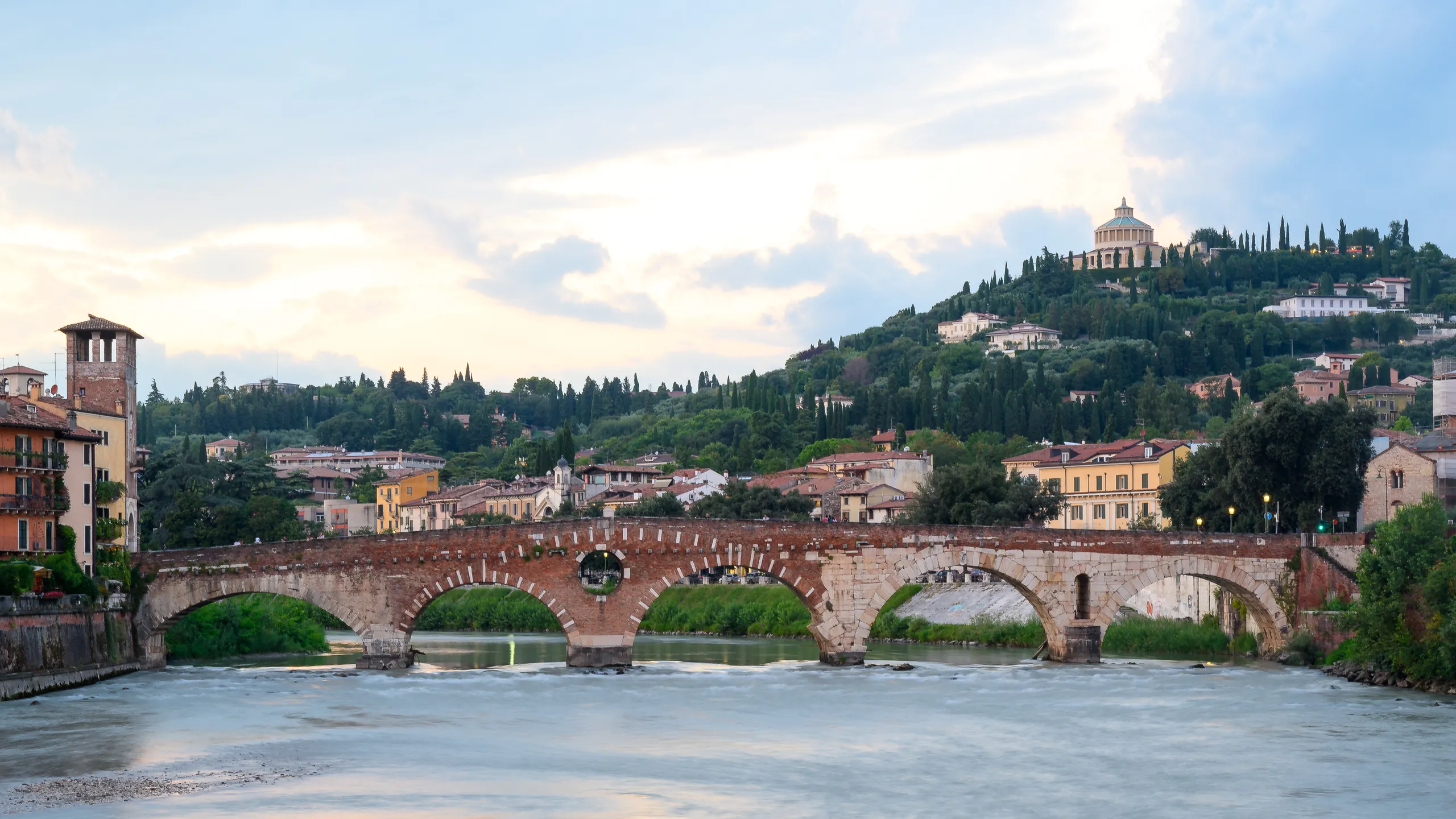 The Roman stone arches of Ponte Pietra crossing a fast river in Verona, with a hill of cypress trees and a sanctuary above.