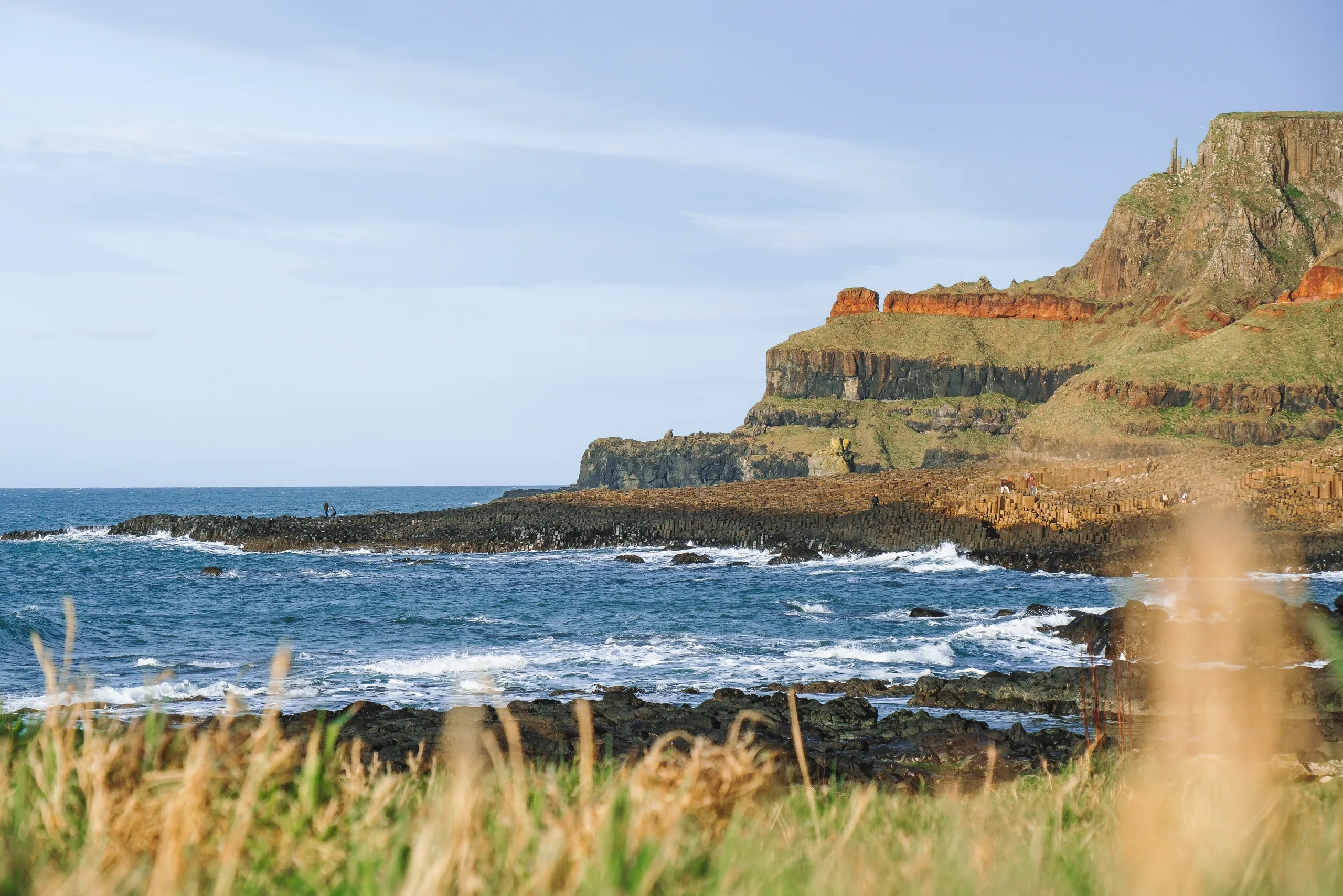 Green-topped basalt cliffs with bands of red rock above a rocky shoreline and blue sea, with golden grass in the foreground.