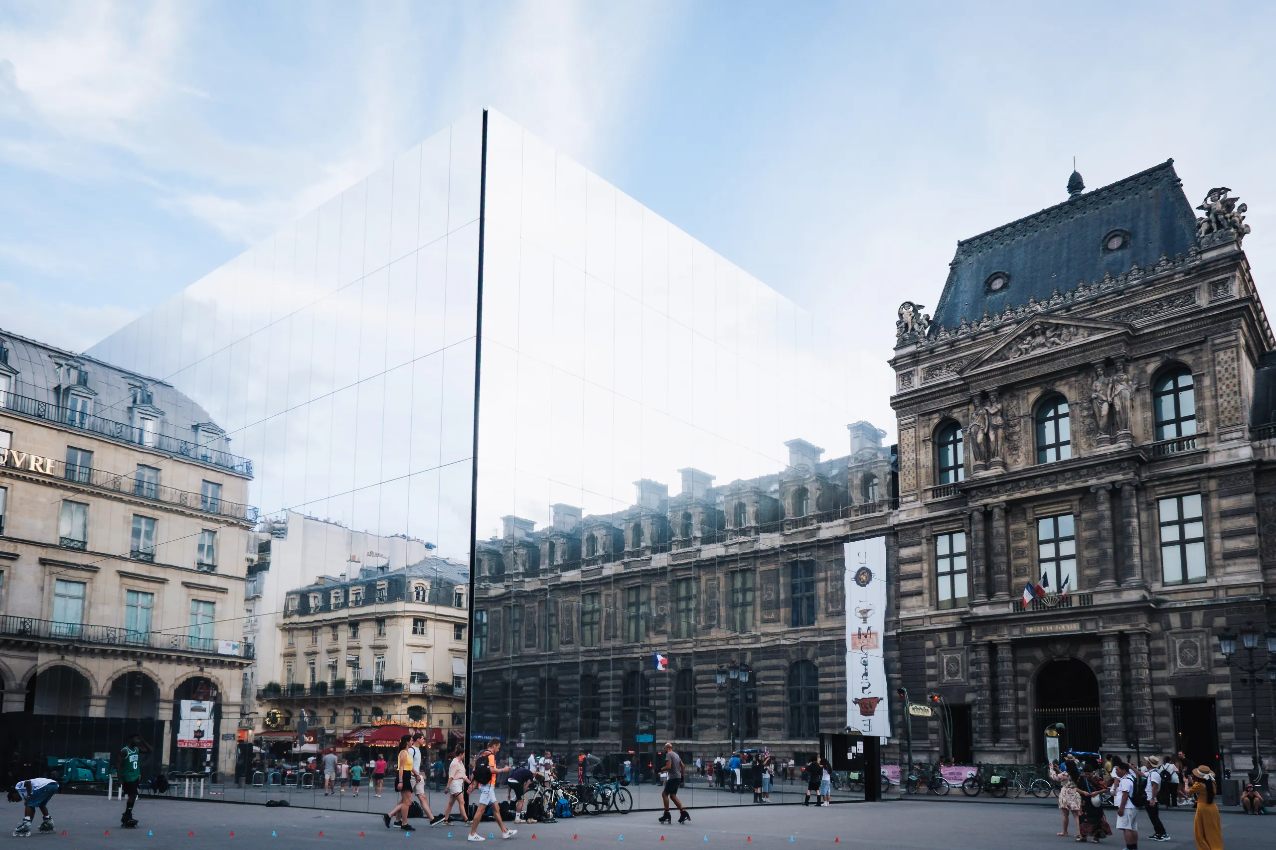 A tall mirrored installation reflecting the sky beside the ornate facade of the Louvre, with people crossing the square in front.