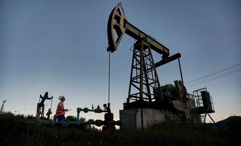Worker in safety gear beside an oil field pump jack at dusk