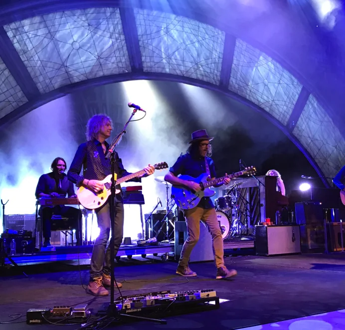 Band playing electric guitars on stage under an arched stained-glass canopy, lit in blue