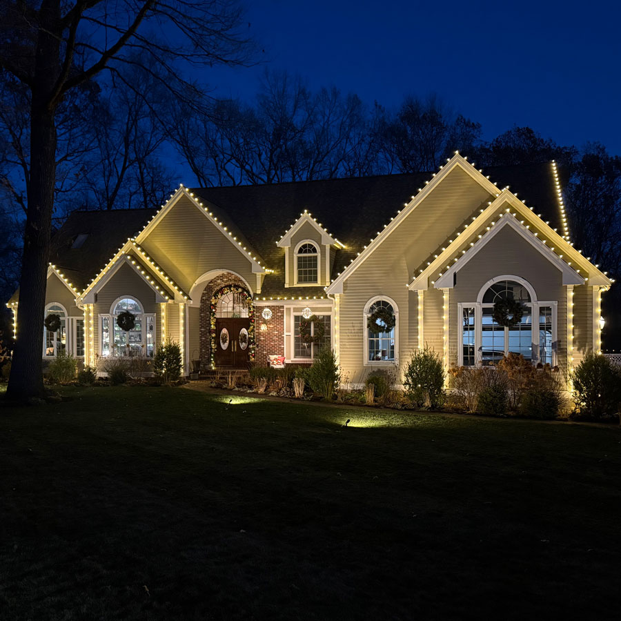 Home at dusk with rooflines and gables outlined in warm white lights and a lit wreath in the front window