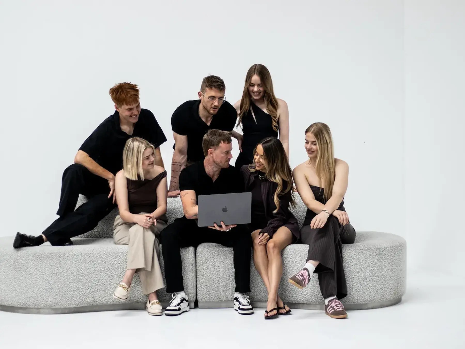Seven young adults sitting and standing on a rounded gray couch looking at a laptop together.