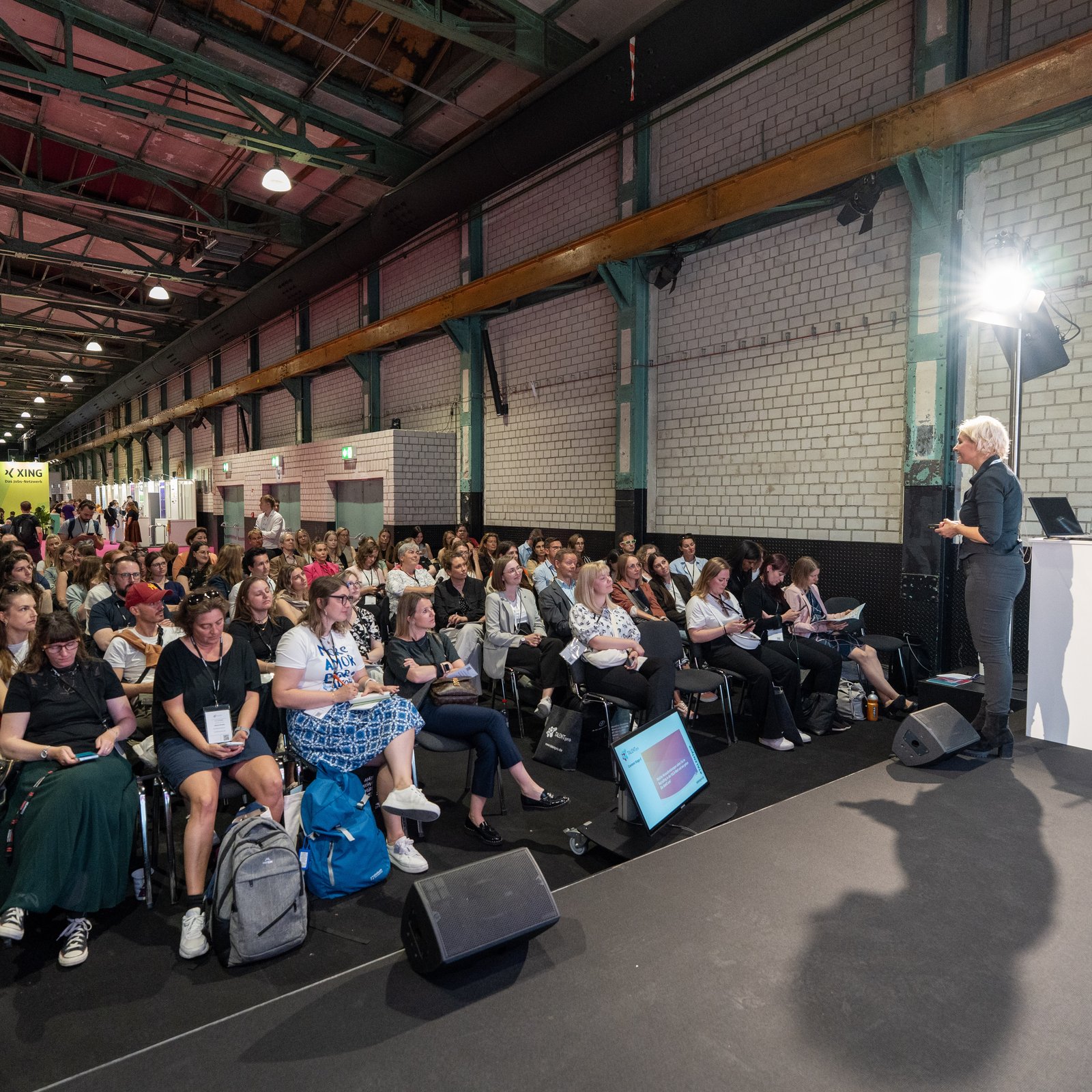 Audience listening to a talk on a stage inside the industrial exhibition hall at TALENTpro 2025 in Munich