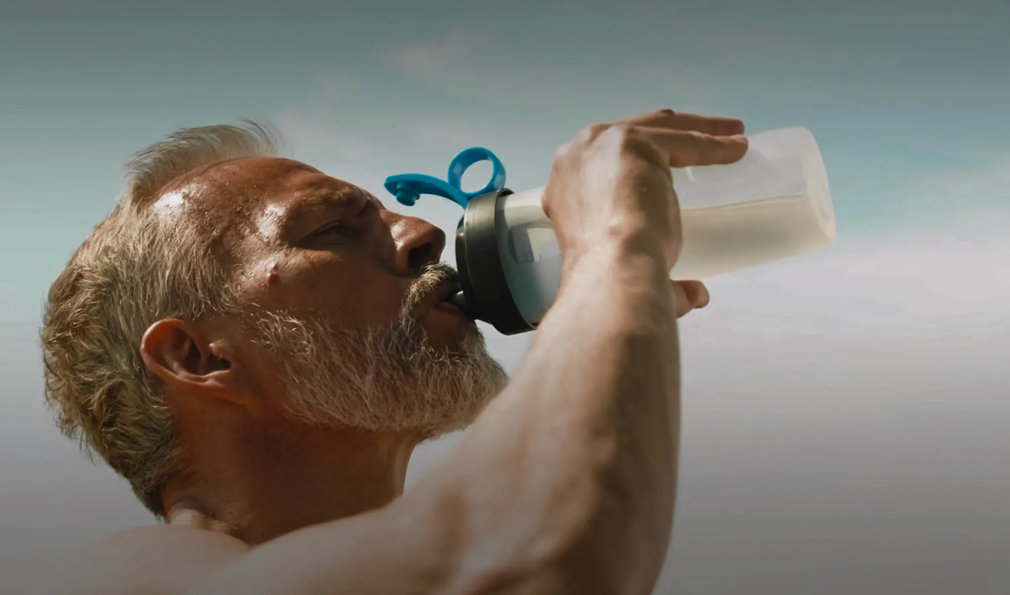 Close-up of a mature man with gray hair and beard drinking from a clear water bottle with a blue flip-top lid against a clear sky.