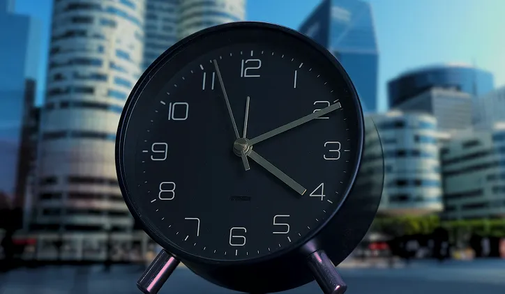 Black analog clock showing time as 11:21 against a blurred background of modern city buildings.