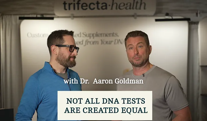Two men stand in front of a Trifecta Health banner, one wearing glasses and a blue jacket, the other in a gray shirt, with the text: 'with Dr. Aaron Goldman' and 'NOT ALL DNA TESTS ARE CREATED EQUAL.'