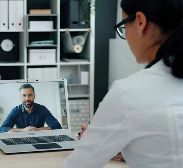 A woman wearing glasses and a white coat is sitting at a desk, participating in a video call with a man displayed on her laptop screen, set in a modern office environment with shelves in the background.