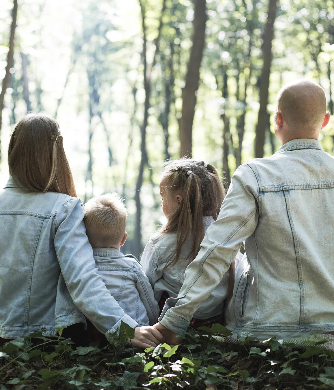 Back view of a family of four sitting in a forest, holding hands, all wearing light denim jackets, surrounded by greenery and tall trees.