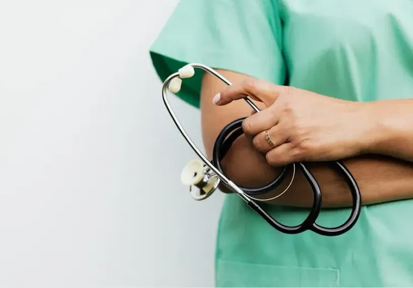 Healthcare professional wearing green scrubs holding a black stethoscope with arms crossed against a plain white background.