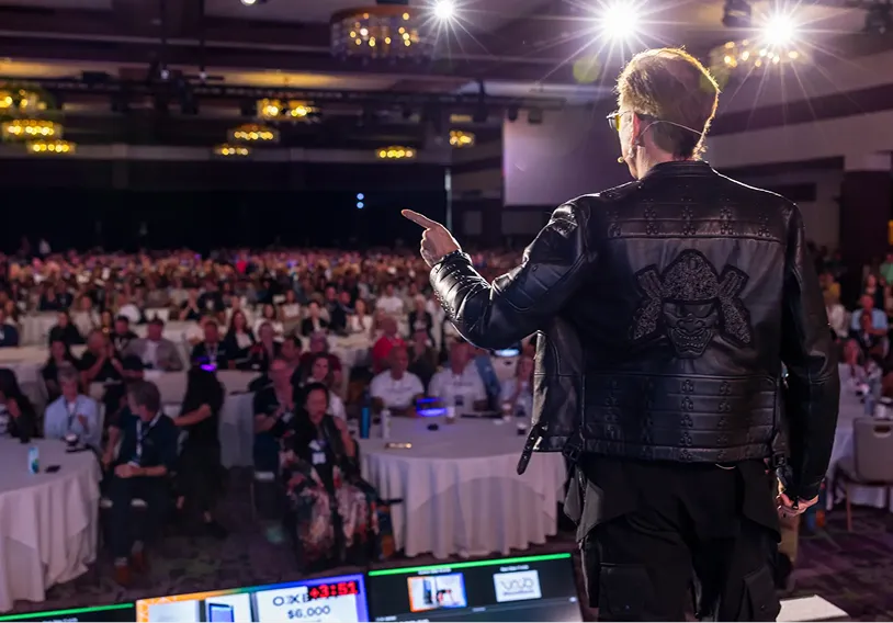 Speaker in a black leather jacket with a samurai helmet design on the back, addressing a large audience seated at round tables in a conference room with bright overhead lights.