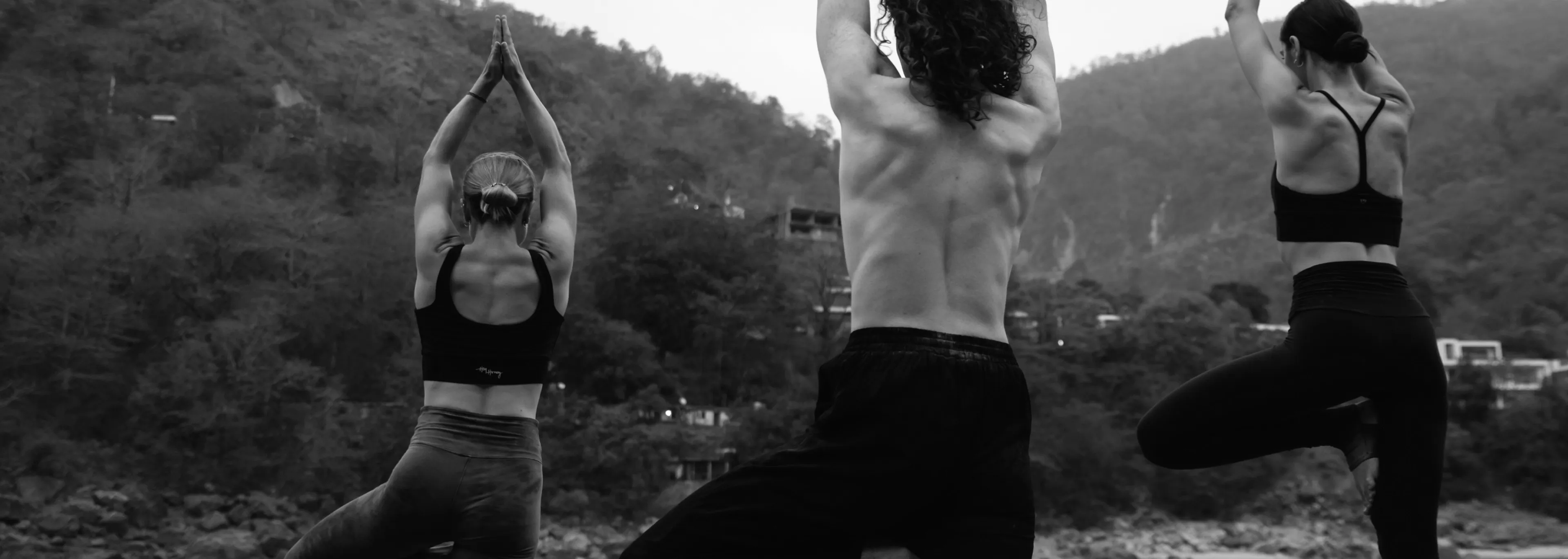 Three people practicing yoga outdoors in a mountainous area, standing on one leg with arms raised above their heads in a tree pose, seen from behind.