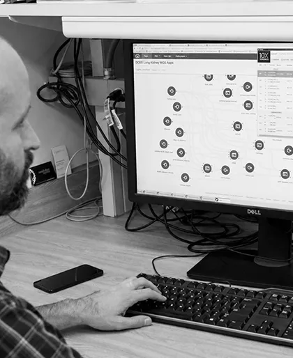 Man with a beard sitting at a desk using a desktop computer with an application showing various circular icons and text on the screen, a smartphone placed on the desk next to the keyboard.