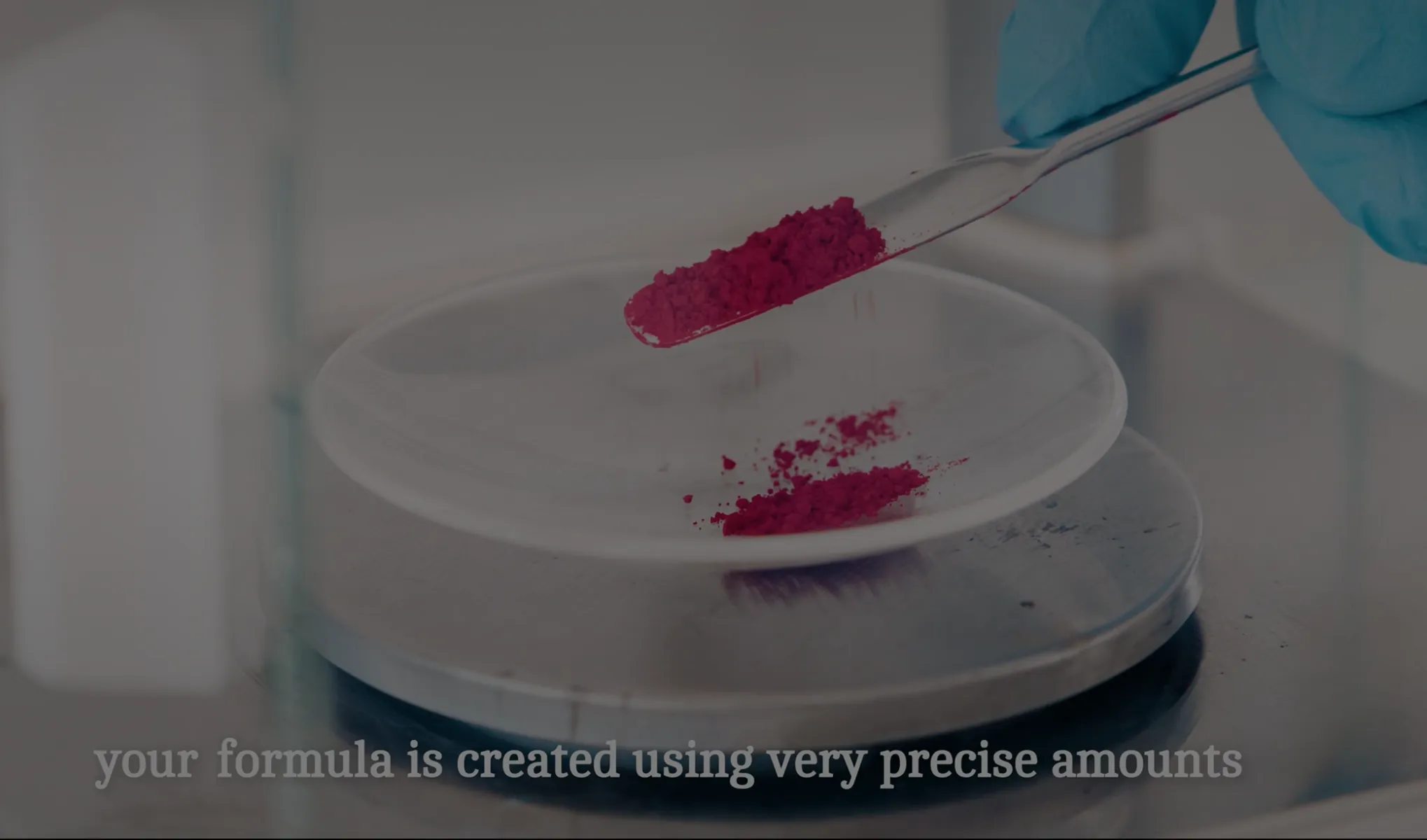 Gloved hand using a spatula to measure and transfer precise amounts of red powder onto a white dish on a digital scale, with text overlay reading 'your formula is created using very precise amounts.'