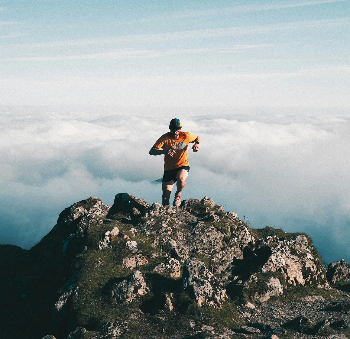 Person wearing an orange shirt, black shorts, and a cap, running uphill on rocky terrain above the clouds during daytime.