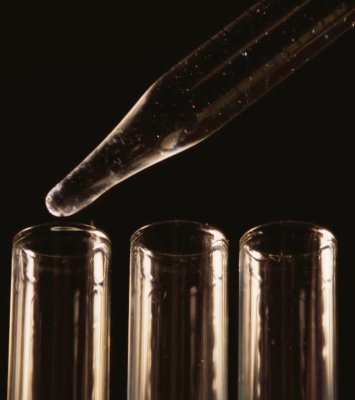 A close-up of a pipette with a drop at its tip positioned above three empty glass test tubes against a black background.