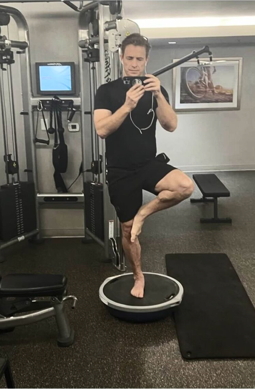 Man in black workout clothes balances barefoot on one leg atop a balance trainer in a gym, taking a mirror selfie with his phone, surrounded by exercise equipment and a bench.