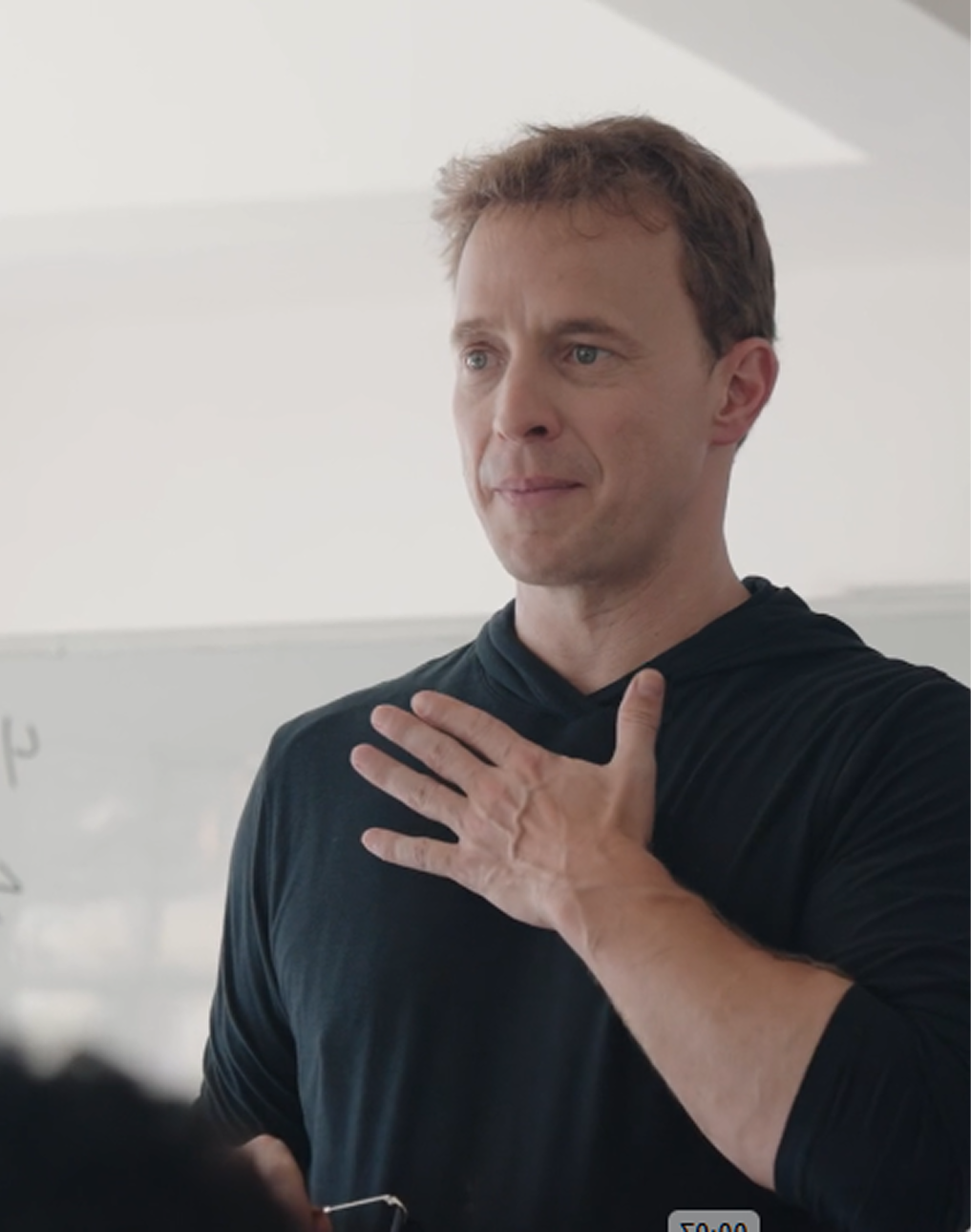 A man with short brown hair wearing a black hoodie places his right hand on his chest while looking slightly to the side with a thoughtful expression in a light-colored room.