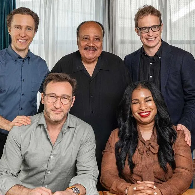 Five people smiling and posing indoors in front of a window with white curtains; three men standing behind two seated individuals—a man with glasses in a gray shirt and a woman with long black hair in a brown blouse.