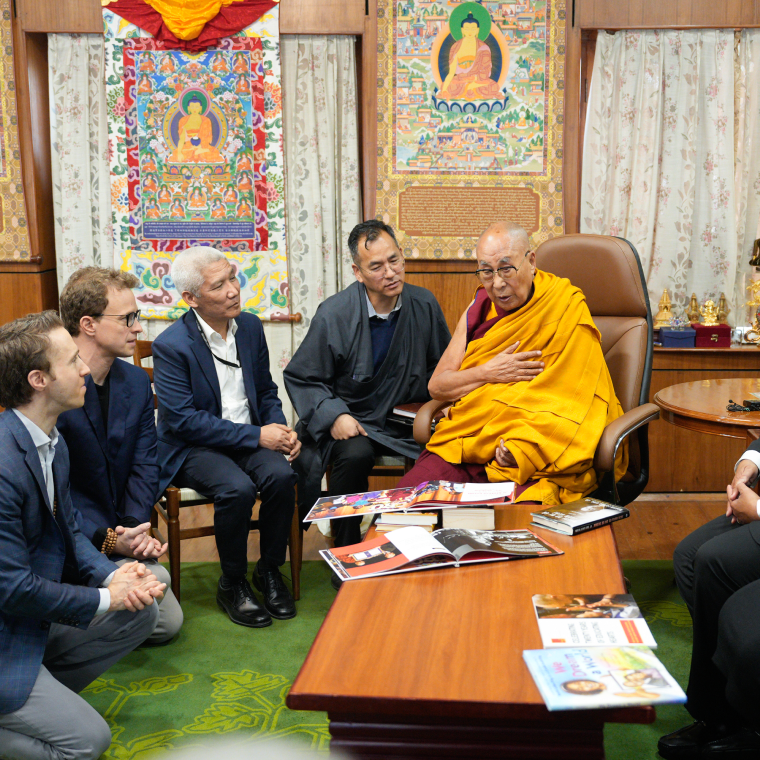 The Dalai Lama seated in a chair wearing a yellow robe, speaking to a group of men seated around a wooden table with books and magazines, in a room decorated with colorful Buddhist paintings and curtains.