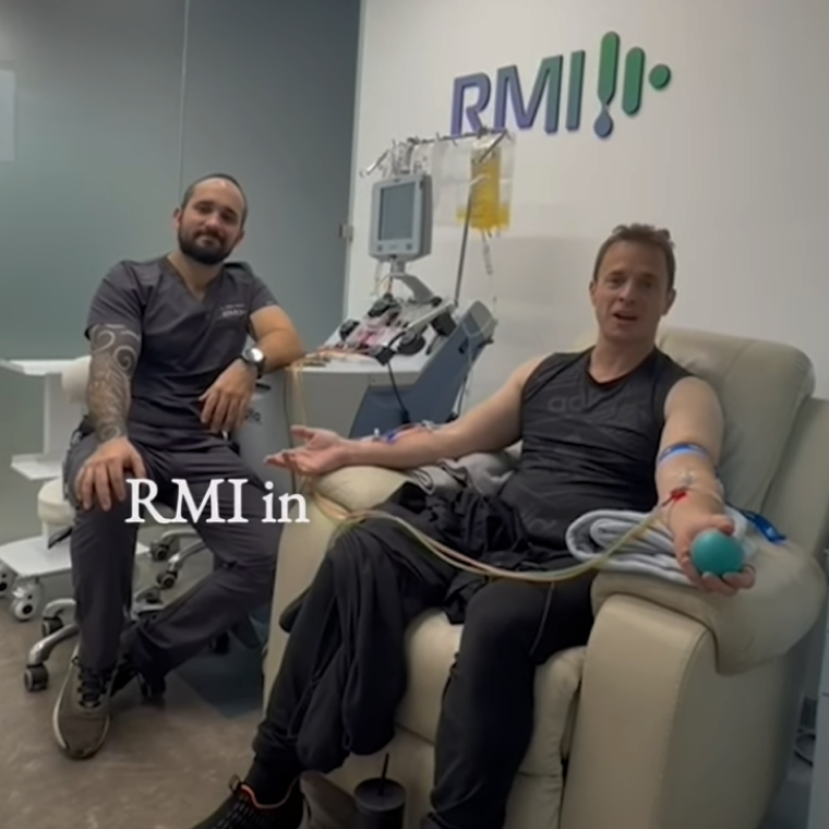 Man donating blood while seated in a medical chair, with tubes connected to an arm and a blue stress ball in hand, accompanied by a healthcare worker in scrubs sitting nearby in a clinical setting with RMI logo on the wall.