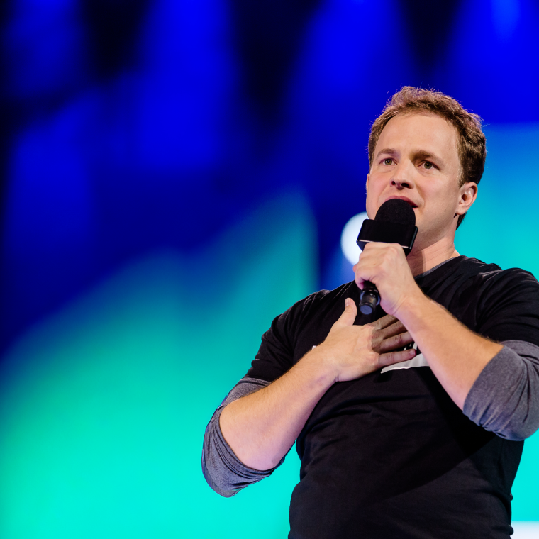 Man with light brown hair wearing a black and grey long sleeve shirt speaking into a microphone with one hand on his chest against a blue and green blurred background.