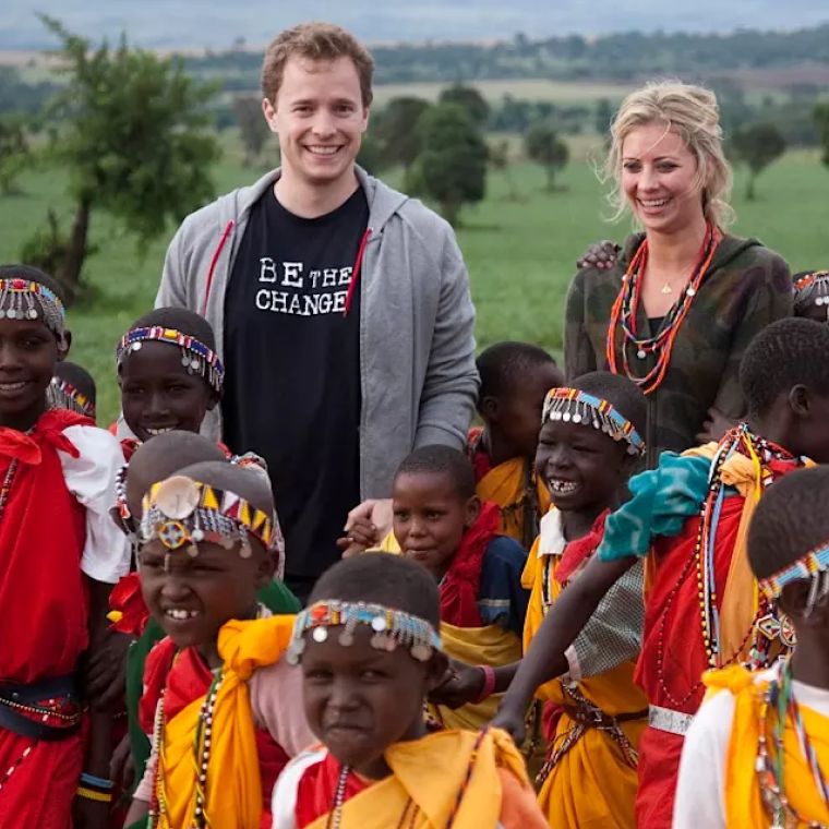 A smiling young man wearing a 'Be the Change' shirt and a young woman with beads around her neck stand outdoors surrounded by smiling children dressed in colorful traditional attire and beadwork, with a green landscape in the background.