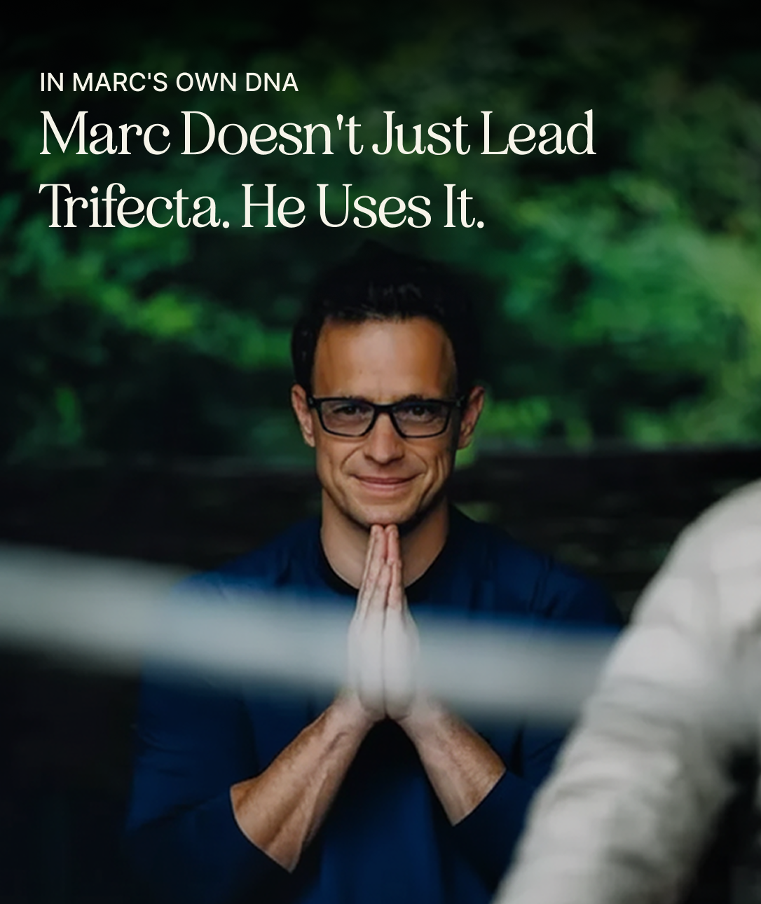 Man wearing glasses and a dark blue long-sleeve shirt, standing with his hands pressed together in a prayer-like position, smiling slightly, with a blurred natural green background and text above him that reads, 'IN MARC'S OWN DNA Marc Doesn't Just Lead Trifecta. He Uses It.'