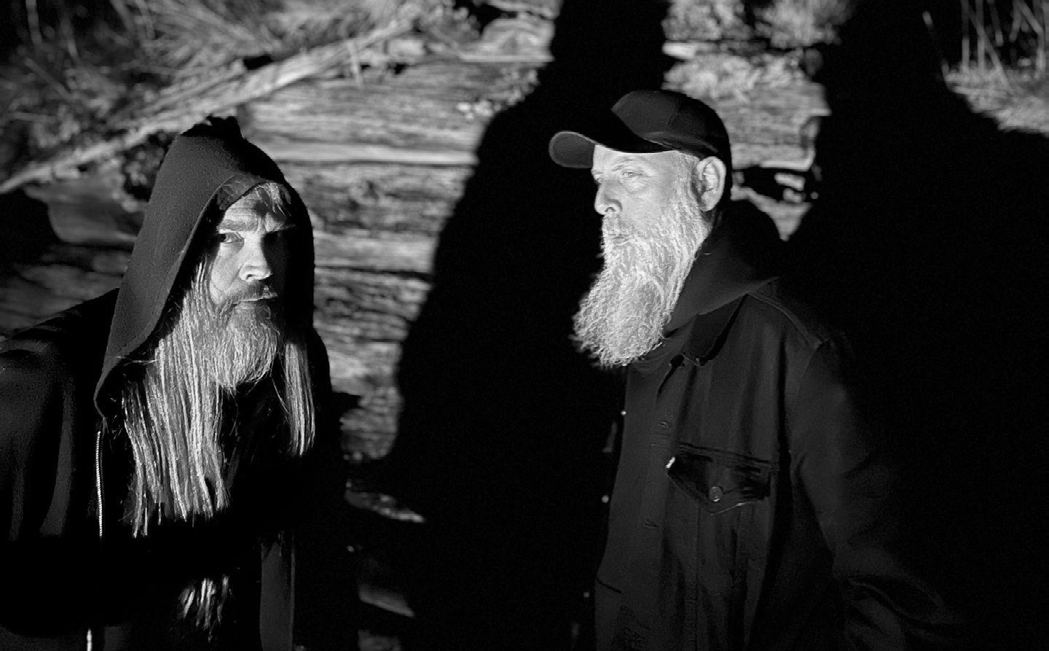 Two band members in dark clothes by a rock wall