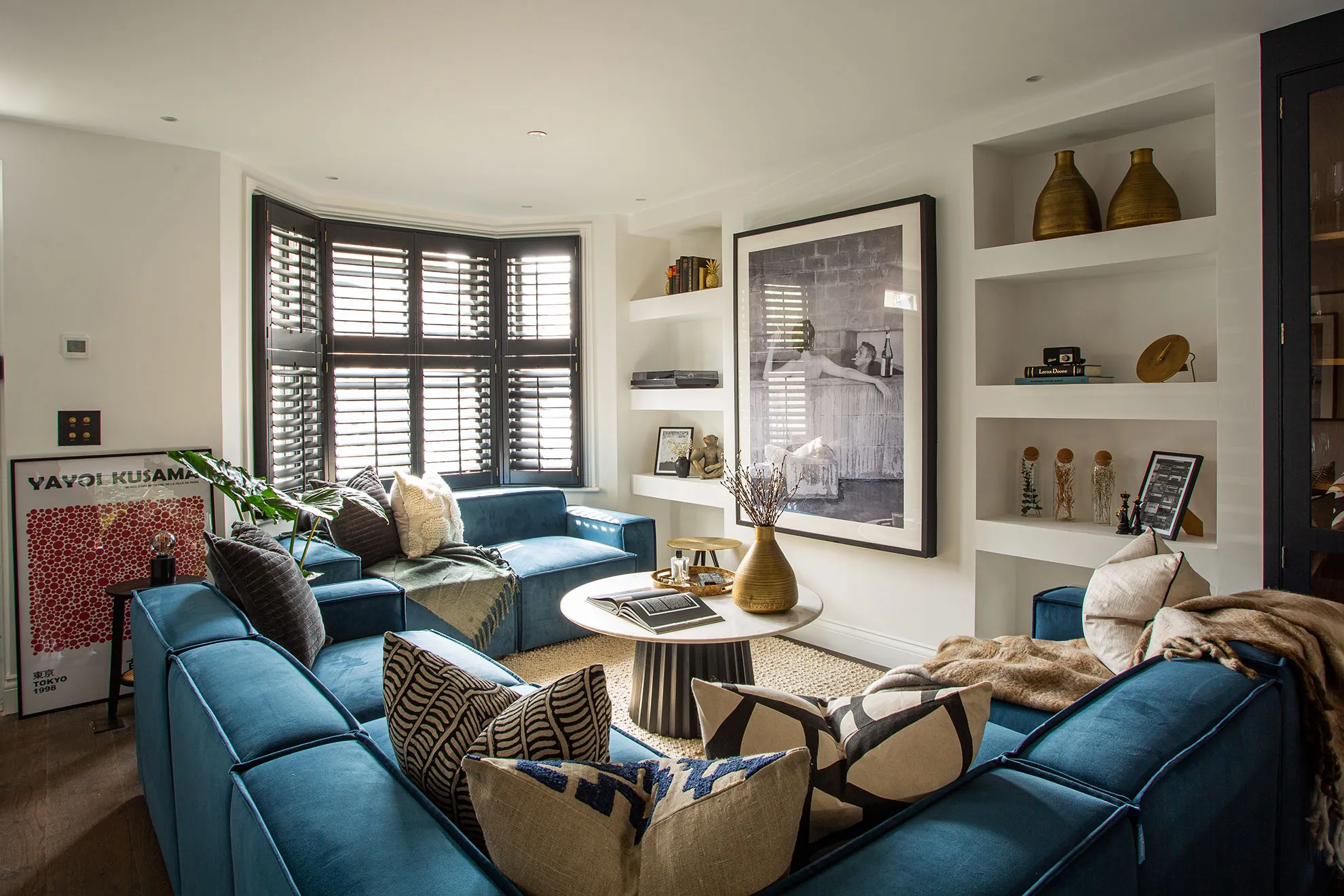 Living room with blue velvet sofas, shuttered bay window and built-in shelving