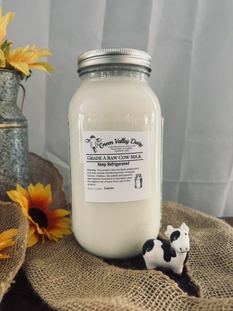 Half-gallon jar of Green Valley Dairy Grade A raw cow milk on burlap beside a cow figurine and sunflowers