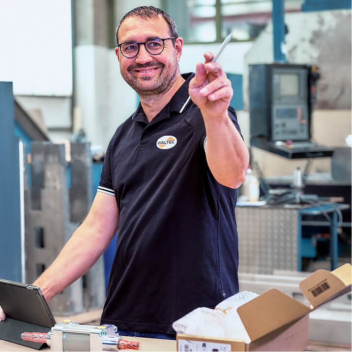 WALTEC After-Sales-Service employee in a polo shirt stands at a workbench holding a pen and smiling while facing the camera. On the workbench in front of him are a tablet and a new machine component.