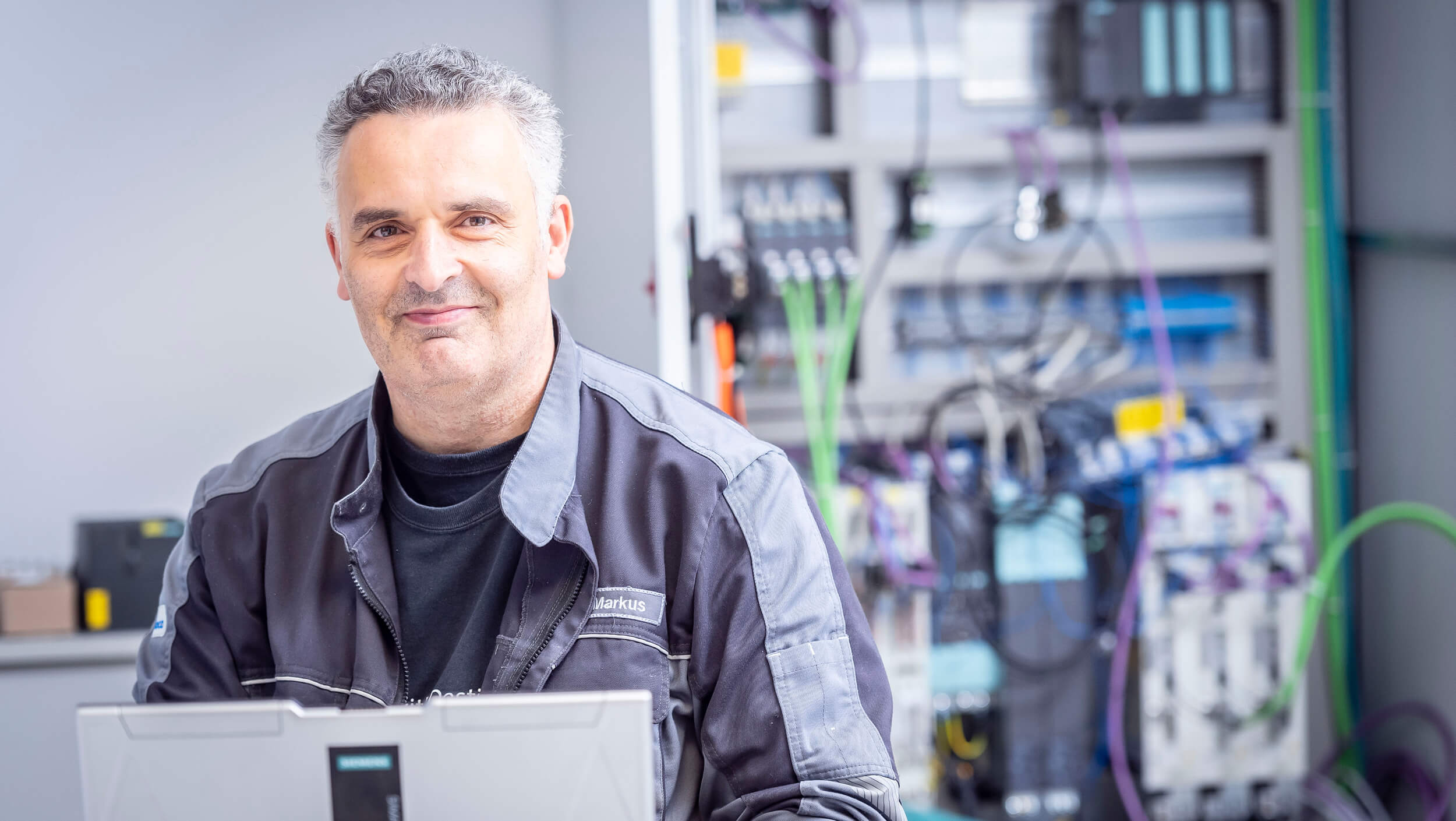 A WALTEC employee in a work uniform sits in front of an open laptop with industrial control panels in the background.