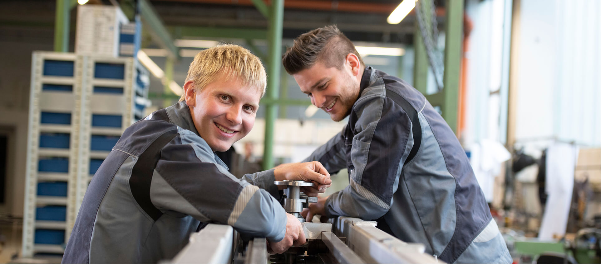 Two WALTEC apprentices in work jackets assemble a metal machine part in an industrial workshop.