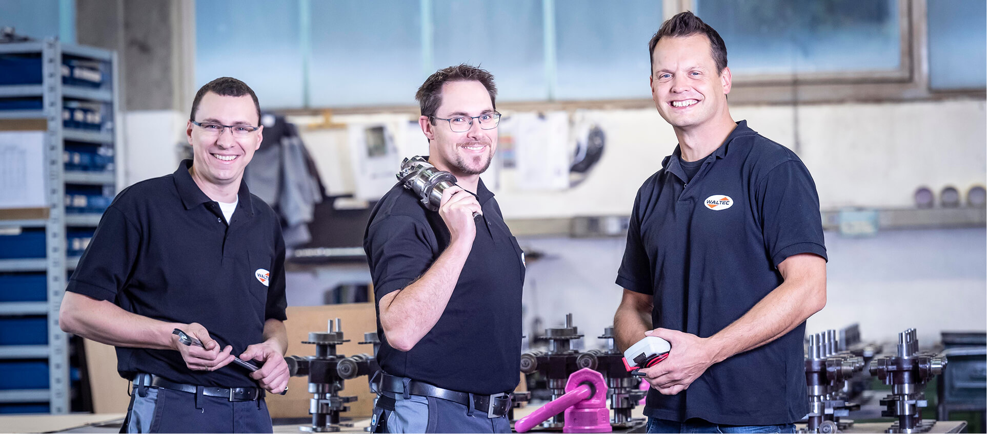 Three WALTEC employees in short-sleeve shirts stand in a workshop holding tools and smiling at the camera