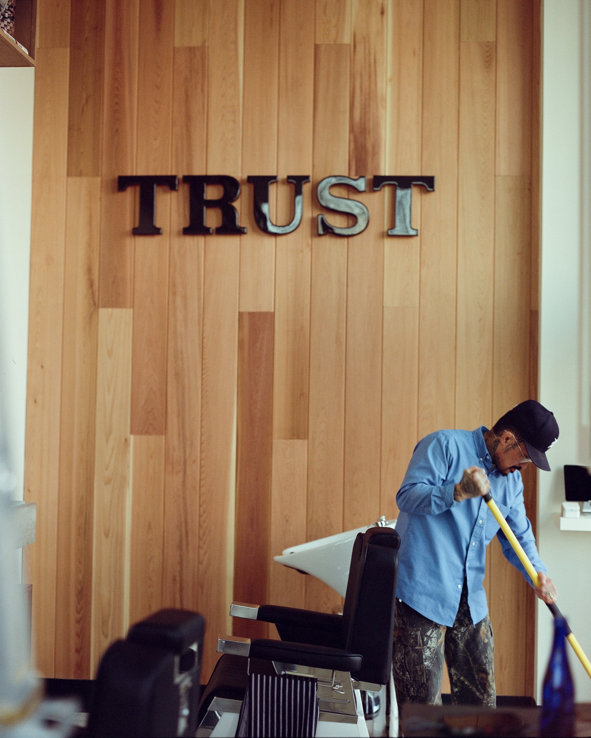 A barber sweeping the floor beneath the TRUST wordmark on the wood wall