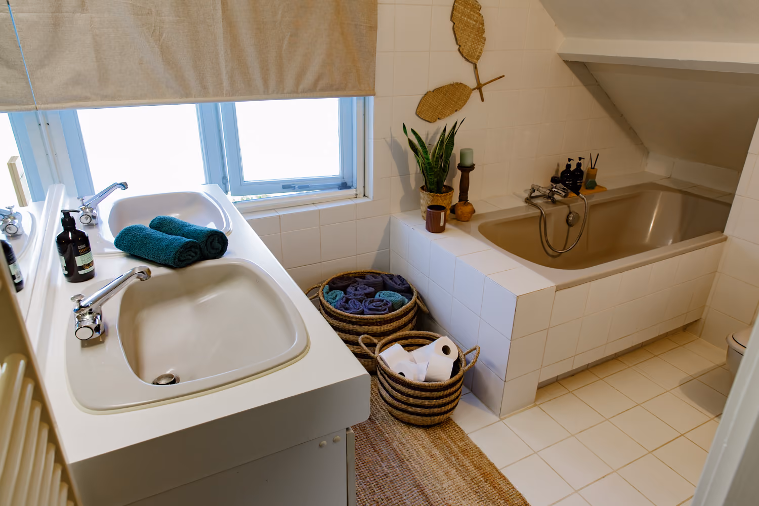 Shared bathroom upstairs with double sink, rolled teal towels, woven baskets and a tiled bathtub under a sloped ceiling